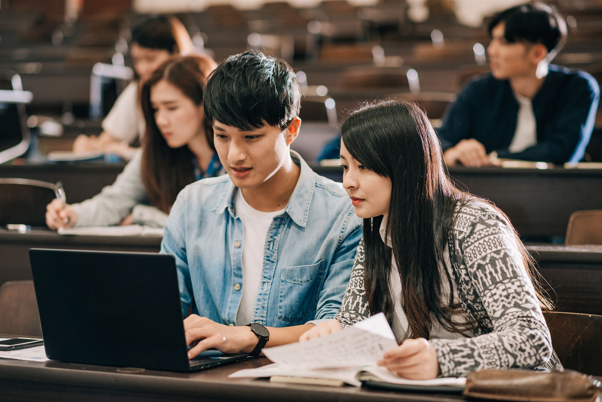 People-seated-on-tiered-chairs-in-lecture-hall,-looking-at-laptops-and-notes
